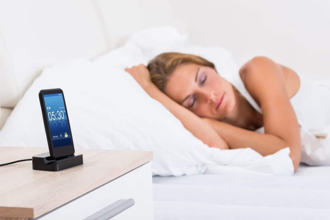 Young Woman Sleeping In Bed With Alarm On Mobile Phone Display
