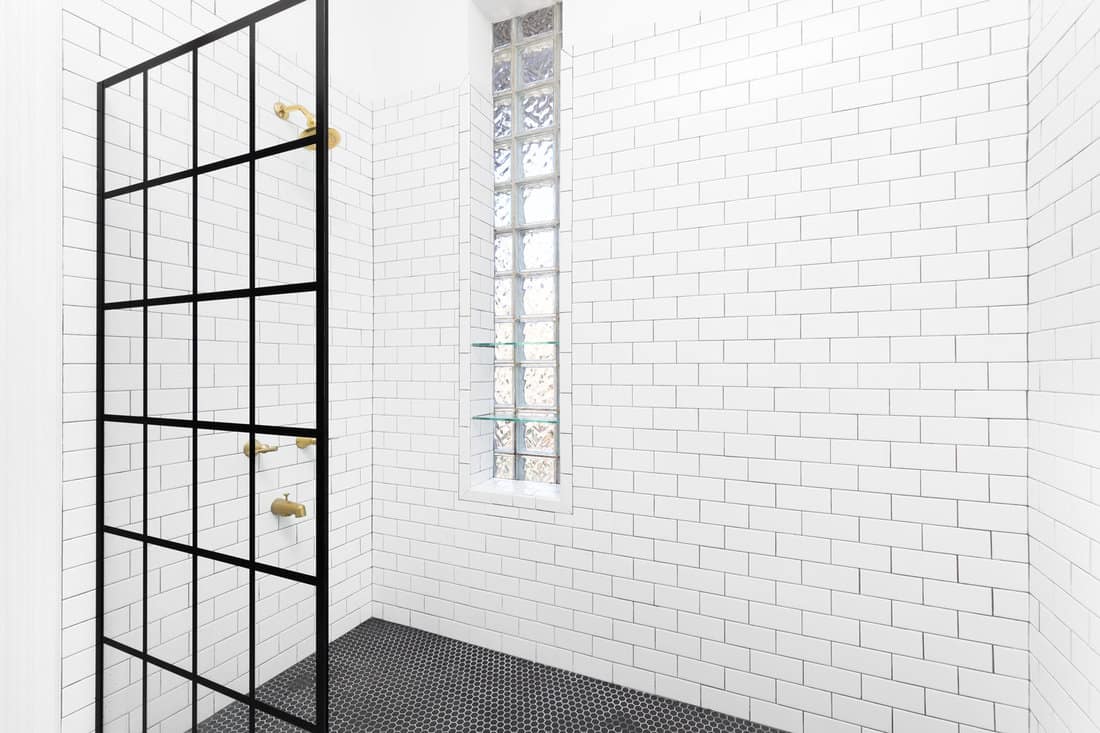 A black French Linea Toulon frameless shower screen with white subway tiles on the walls, black circular tiles on the floor, and glass block windows with shelves.