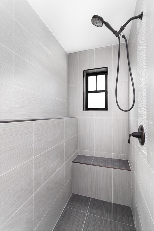 A luxury walk-in shower with tiled walls and bench, a black framed window, and black shower head.