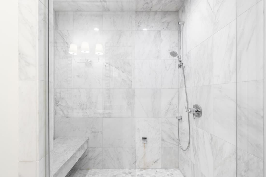 A shower with white marble tile and glass walls, a marble hexagon tiled floor, bench seat, and chrome showerhead.