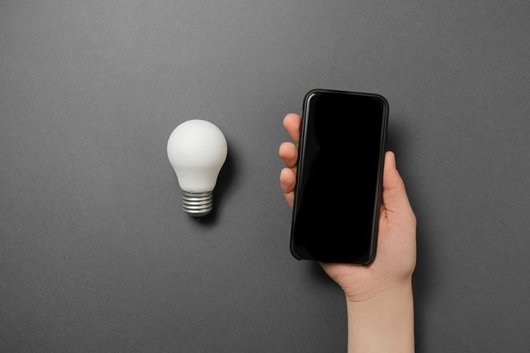 Creative top view flat lay of light bulb and woman holding modern smartphone mockup composition with copy space on grey background, How To Group Smart Lights [Step By Step Guide]