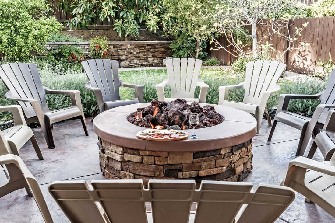 Large outdoor fire pit surrounded by wooden rocking chairs, beautifully landscaped backyard, with the glass of wine and food platter. 