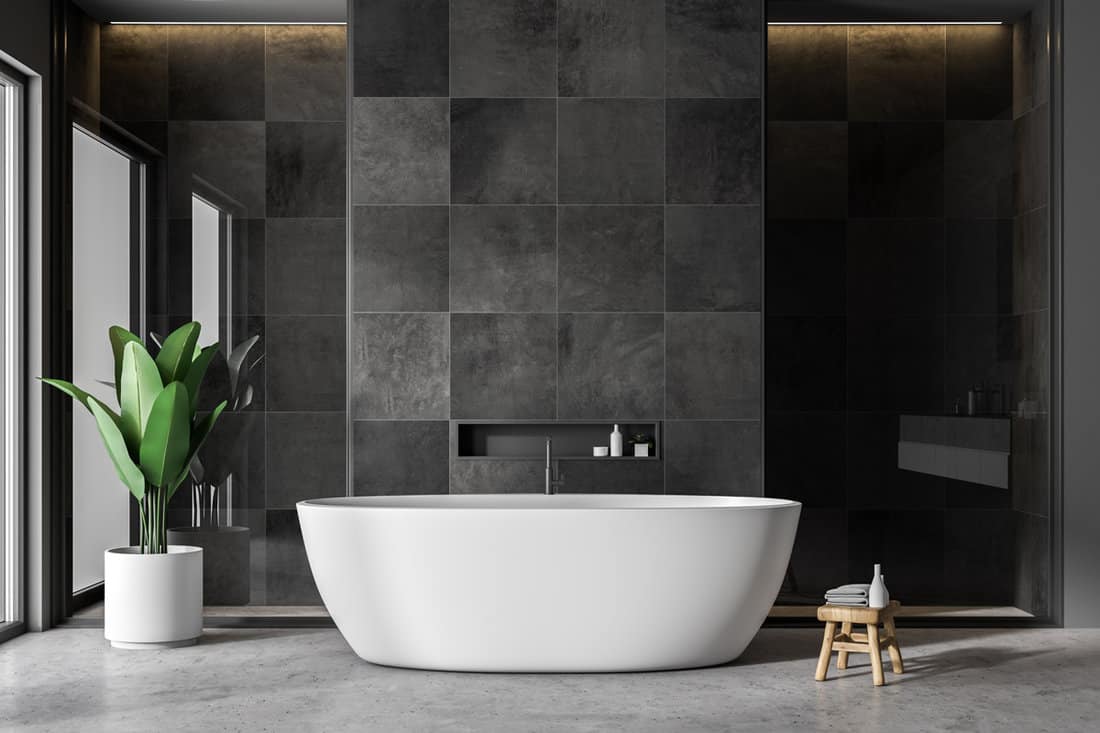 Modern bathroom interior with black tile walls, concrete floor and white bathtub.
