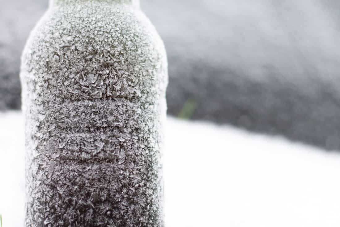 Partially blurred bottle of beer covered with hoarfrost in snow on dark background.