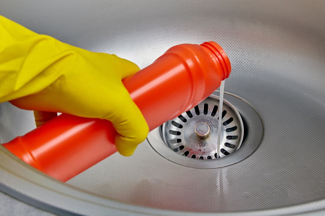 Person's hand in a yellow rubber glove pours pipe cleaner down the drain of a metal kitchen sink 