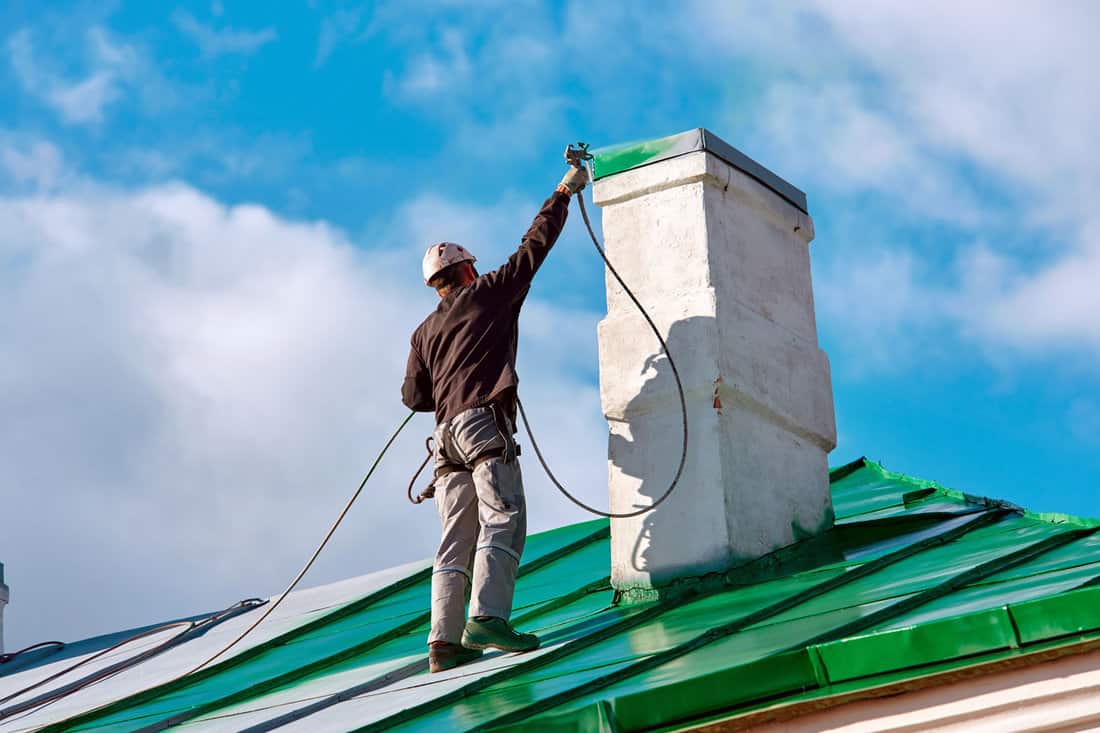 Worker of Industrial Alpinist Services painting chimney on the roof with paint spray gun. Professional climber wearing uniform, helmet and using safety harness. Risky job. Extreme occupation. 
