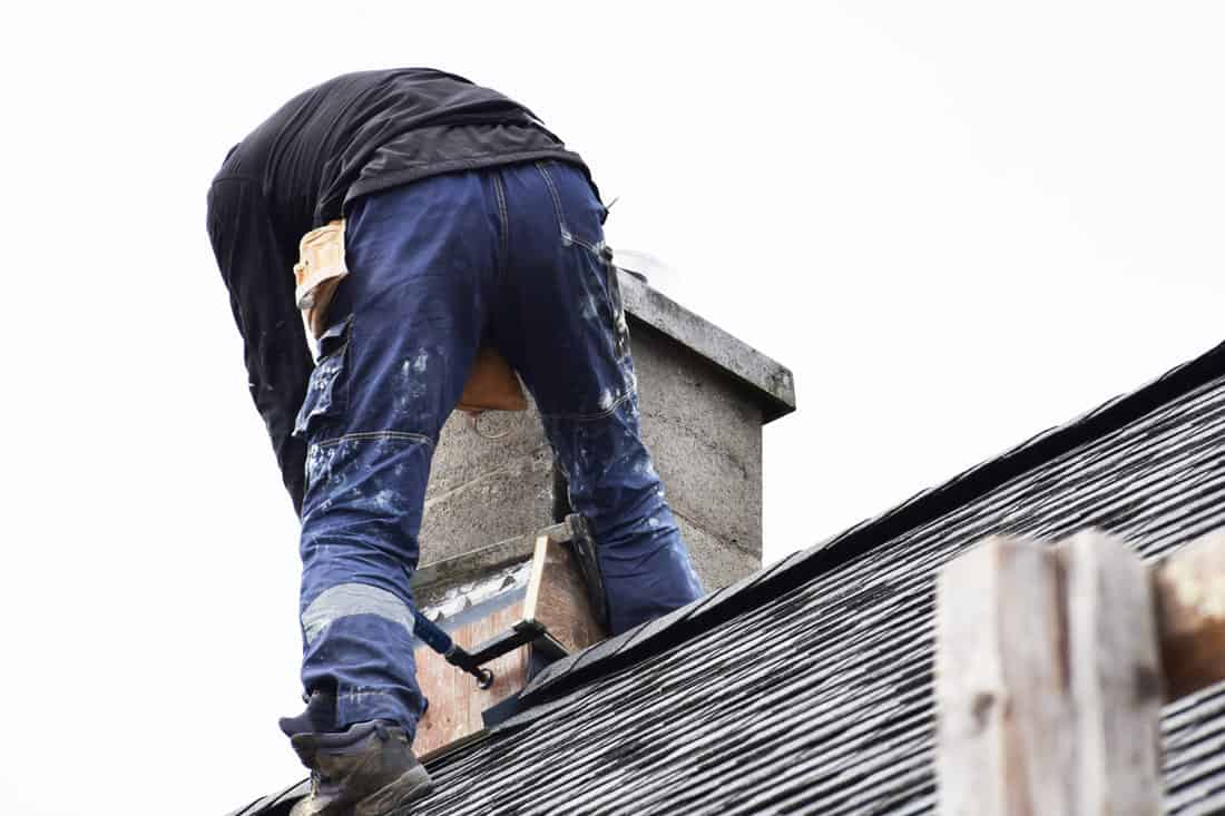 Roofer construction worker repairing chimney on grey slate shingles roof of domestic house, sky background with copy space.