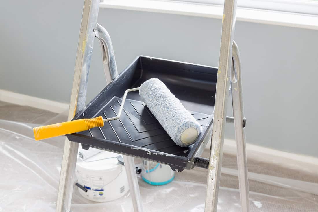 Painting and decorating with a roller and tray on a set of metal step ladders with cans of paint