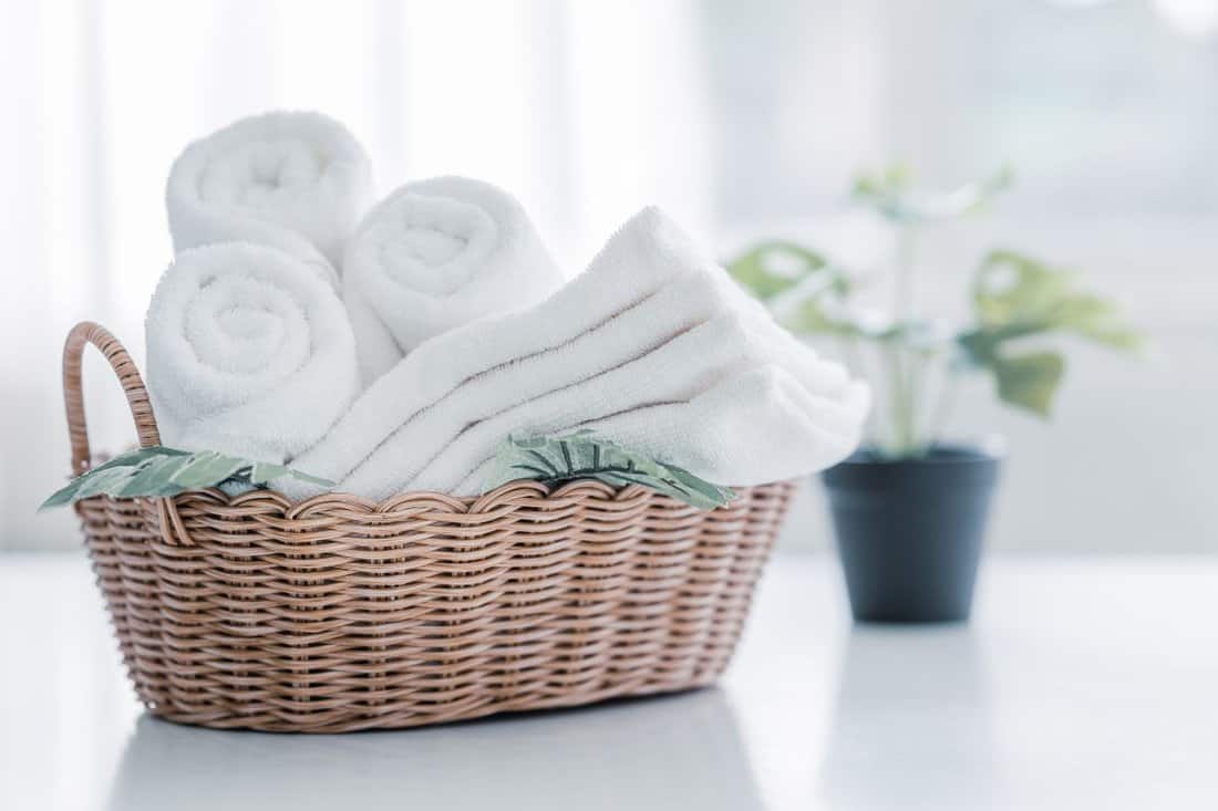 white cotton towels in basket with green plant on white counter table inside a bright bathroom background, copy space for product display montage. 