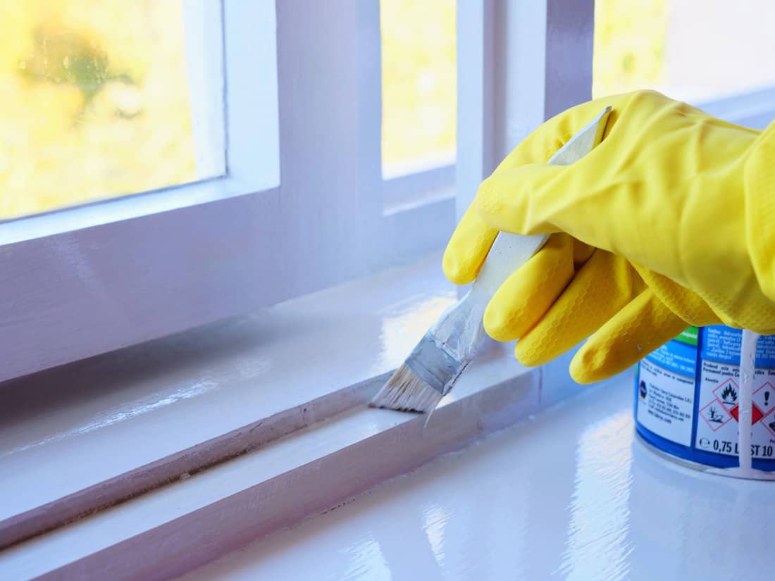 Gloved hand holding a paintbrush painting a window sill with glossy white paint