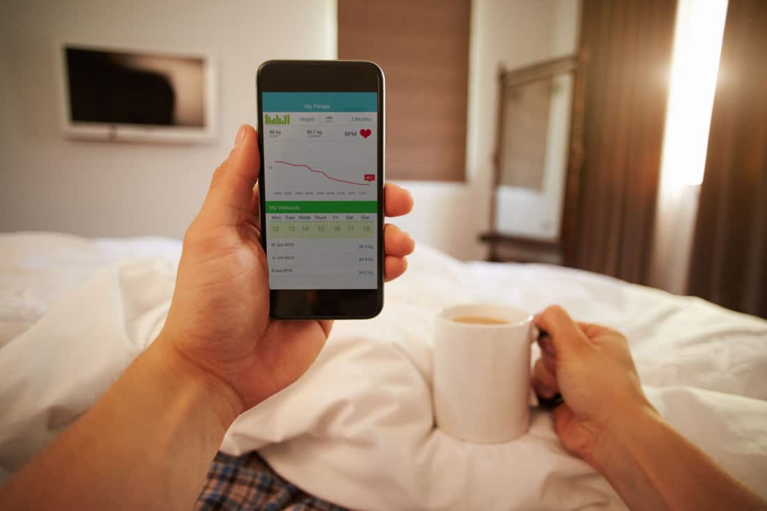 Man In Bed Looking At Health Monitoring App On Mobile Phone