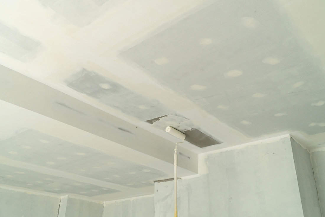 A photograph captures the painting process of a recently installed ceiling, where a ceiling paint roller is used; the new ceiling is being primed first before receiving two coats of paint.