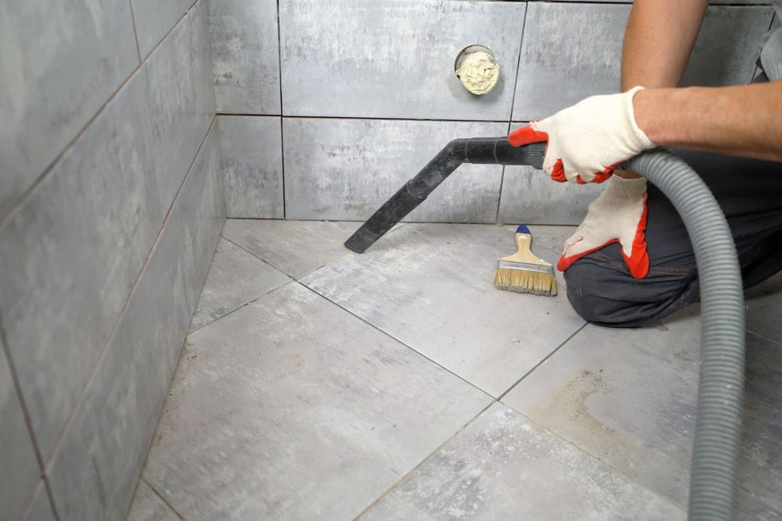 A worker vacuums the floor in a tiling bathroom. A man vacuums ceramic tiles on the floor. Cleaning up debris after laying tiles with a vacuum cleaner. The worker removes the garbage.