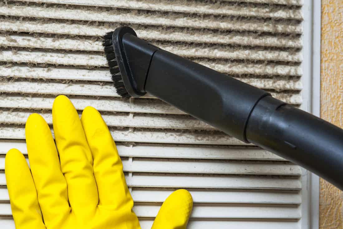 A gloved hand and a vacuum cleaner pipe equipped with a brush work in tandem to remove loose particles from the freshly mudded popcorn ceiling.