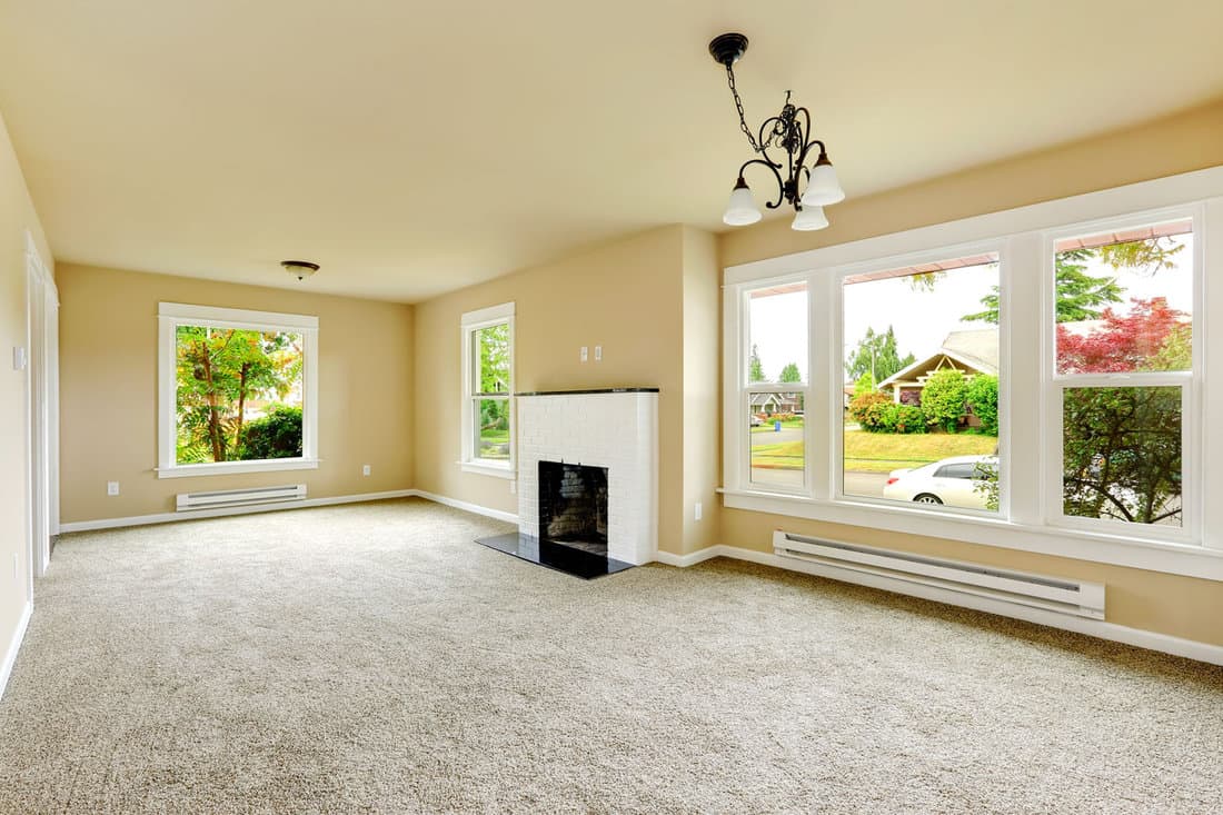 An unfurnished home interior. Family room with ivory walls, light grey carpeted floor, and a white brick fireplace. Frieze carpet accents the space.