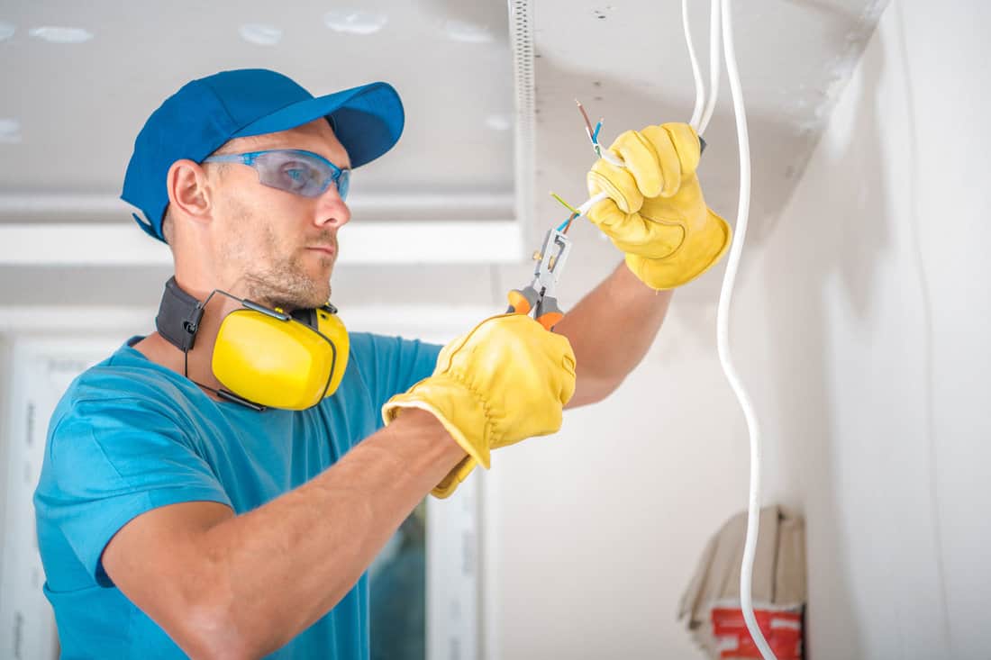 An electrician, wearing protective gloves and glasses, is reconnecting a doorbell.