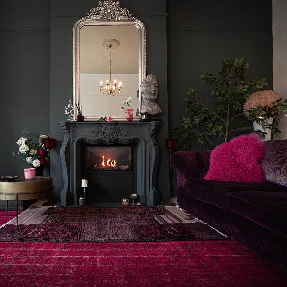 Living room with black  walls and  fireplace. Deep red layered rugs on floor. Burgundy couch with bright pink fluffy throw pillow. Romantic style.