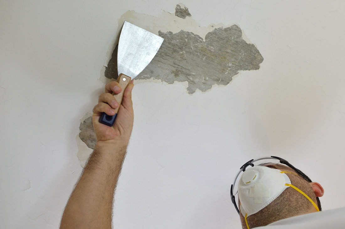 Man scraping a popcorn ceiling with a scraper of putty knife, preparing it for smoothing.
