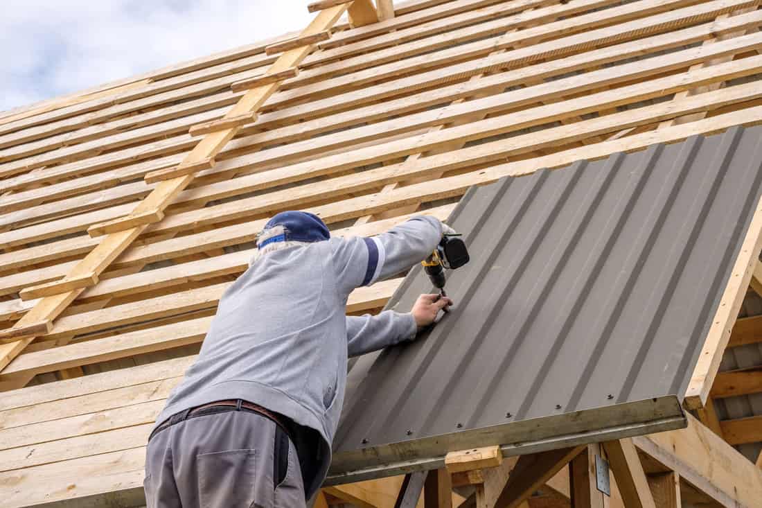 Senior gray-haired Construction man using a screwdriver, fastens a roofing sheet to wooden rafters on the roof of a country house under construction. The physical activity of the elderly.
