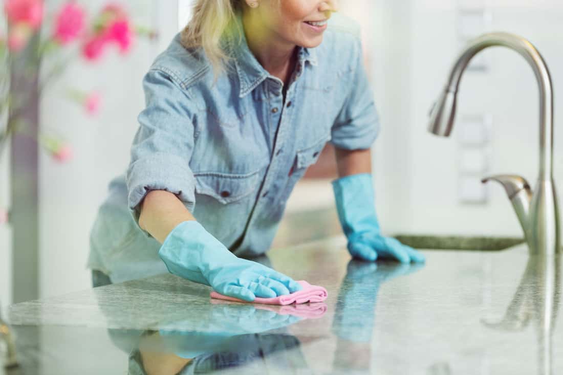 The image captures a woman using a microfiber cloth to expedite the drying process of a countertop recently treated with granite sealer, showcasing her meticulous care for the surface's upkeep.