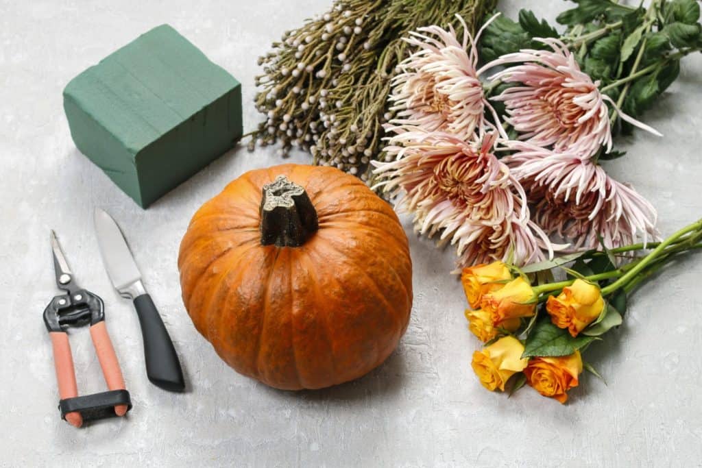 Hand arranging delicate pink flowers and greenery inside a hollowed-out pumpkin with floral tools beside.