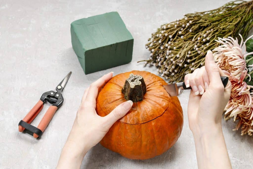 Hands preparing a pumpkin centerpiece with floral foam, shears, and fresh flowers