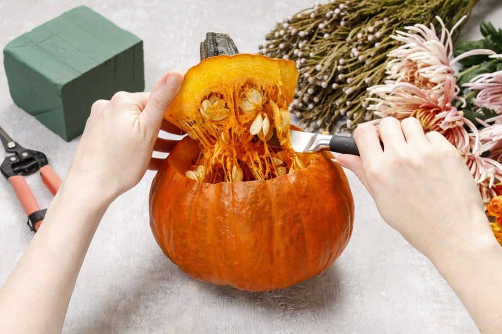 Hands scooping out seeds and pulp from a hollowed-out pumpkin next to floral foam, shears, and assorted flowers