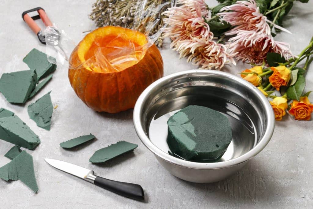 A DIY setting with a hollowed-out pumpkin containing a plastic bag, surrounded by broken pieces of green floral foam, a stainless steel bowl with a piece of wet foam, a floral knife, chrysanthemum flowers, yellow roses, and other crafting items 