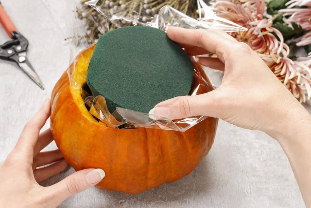 A close-up view of a person's hands placing a green floral foam inside a plastic-lined hollowed-out pumpkin. In the background, there are floral scissors, chrysanthemum flowers, and other crafting items
