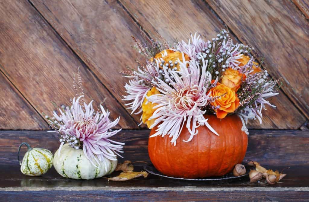 Two decorative pumpkin centerpieces on a rustic wooden surface; one large pumpkin filled with pink and orange flowers and a smaller, white pumpkin with pink floral accents.
