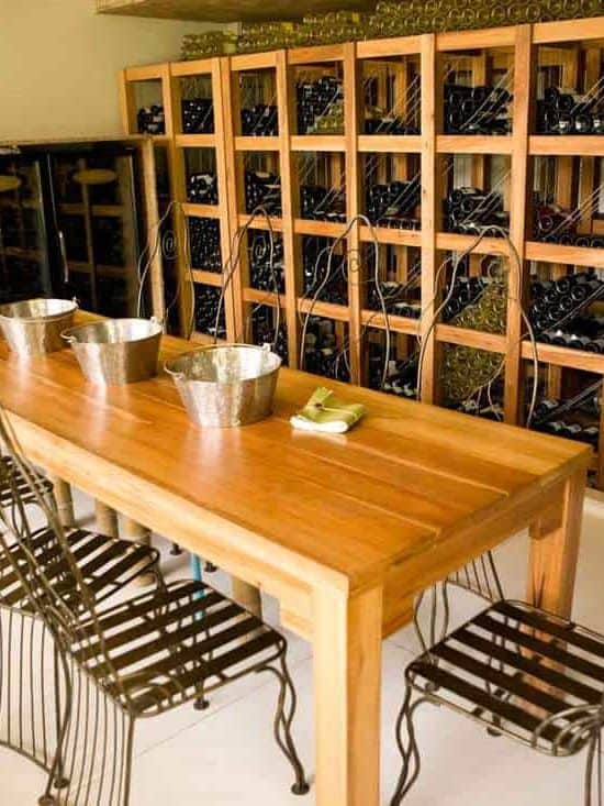 A rustic basement cellar with lots of wine on perfectly arranged wines, a long solid hardwood tables, and metallic chairs on the sides