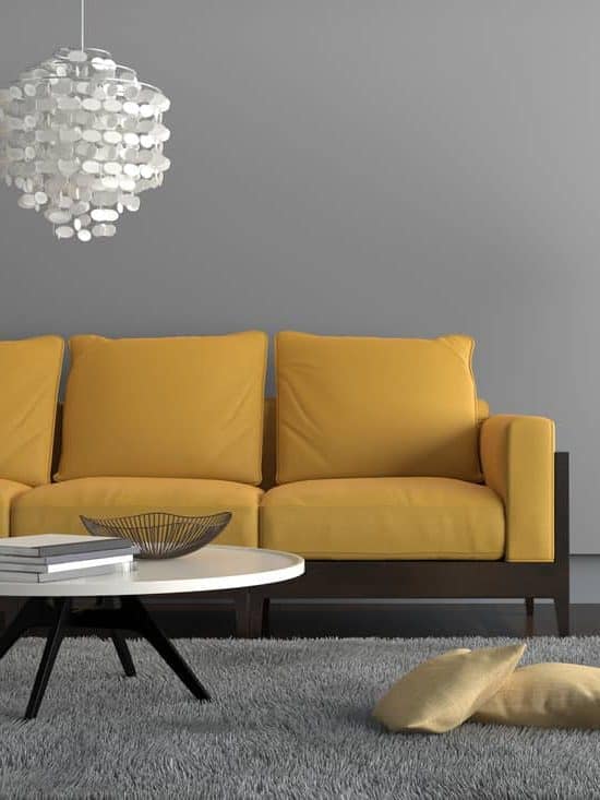  living room showing a yellow sofa, with a lamp, ornate pendant and a table with decoration. There is a gray carpet with two pillows on it. Gray wall surrounding the scene with wooden floor
